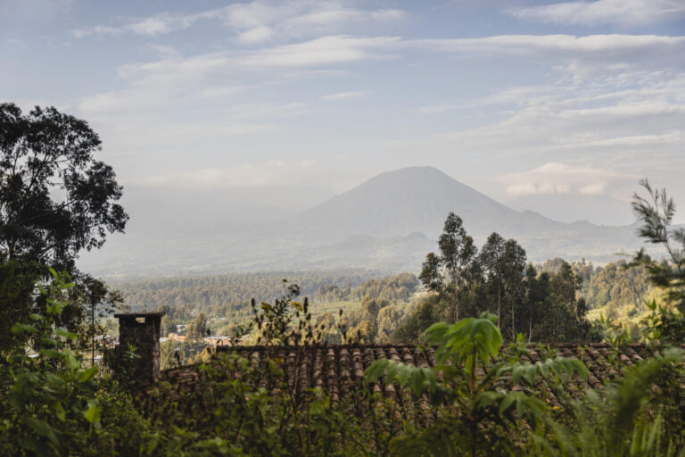 Volcano-shaped mountain rising over a valley, seen from a foreground of trees and a tiled roof.