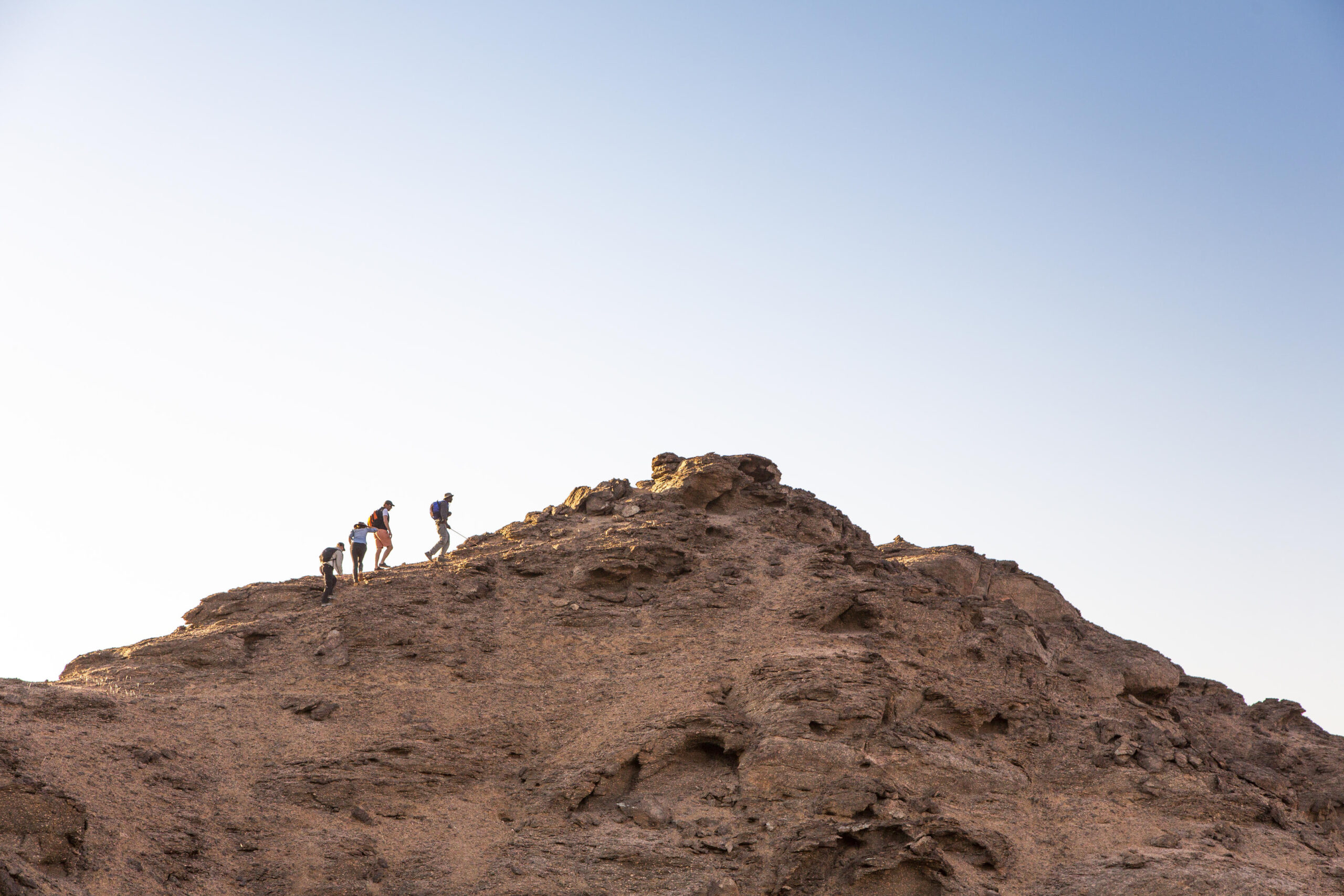 Hikers climb a brown rocky hill against a pale blue sky, each with backpacks and trekking gear.