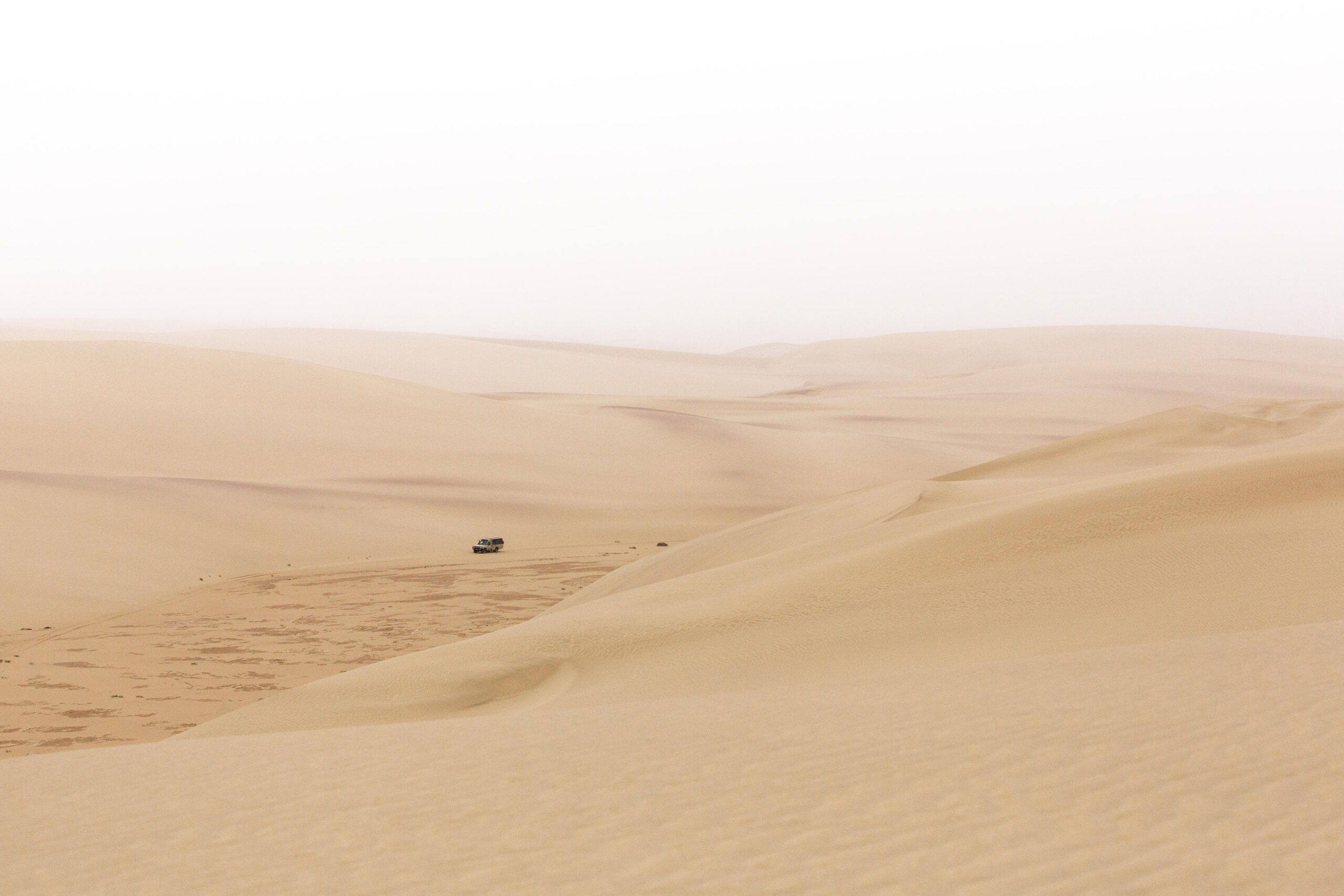 Desert landscape with rolling golden sand dunes and a lone off-road vehicle in the distance.