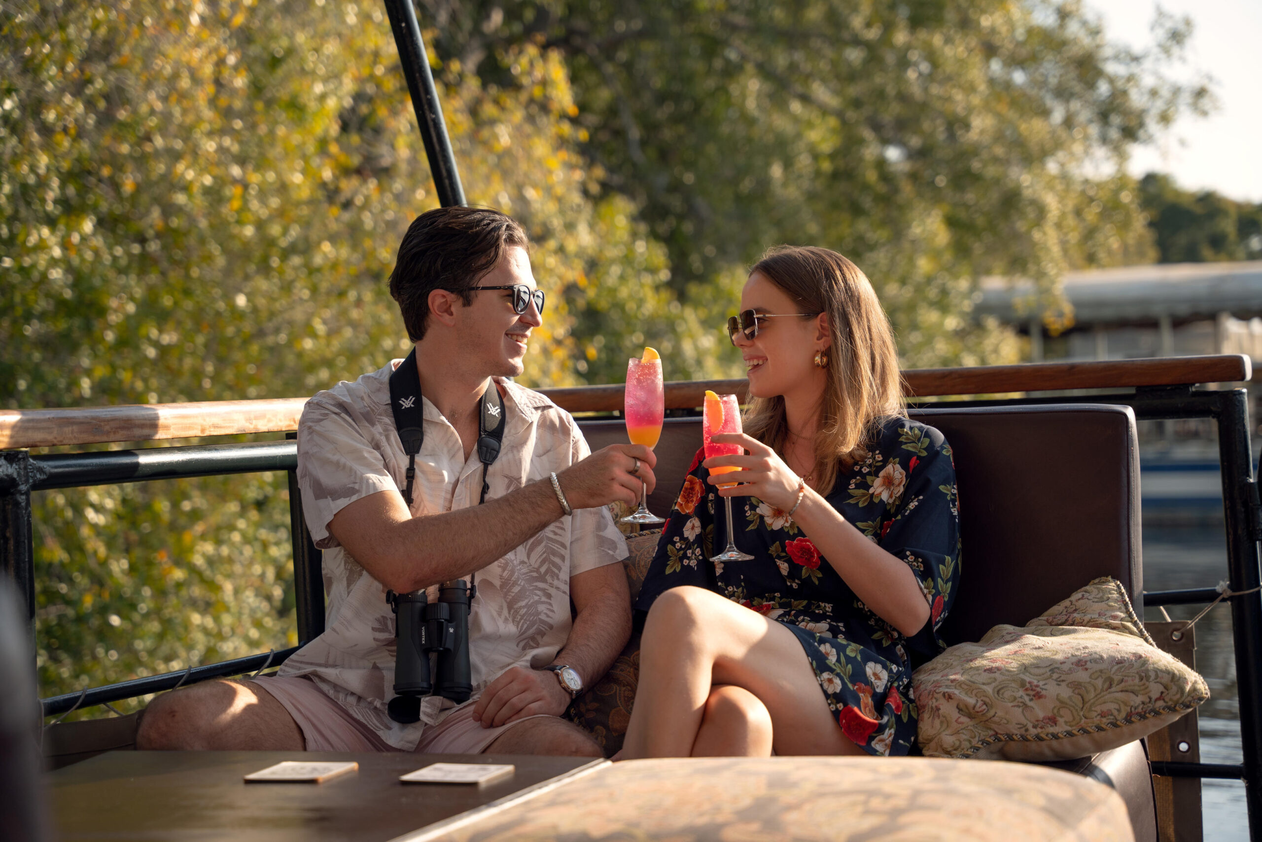 Couple toasting colorful cocktails on a boat deck, wearing sunglasses and summer outfits, with trees in the background