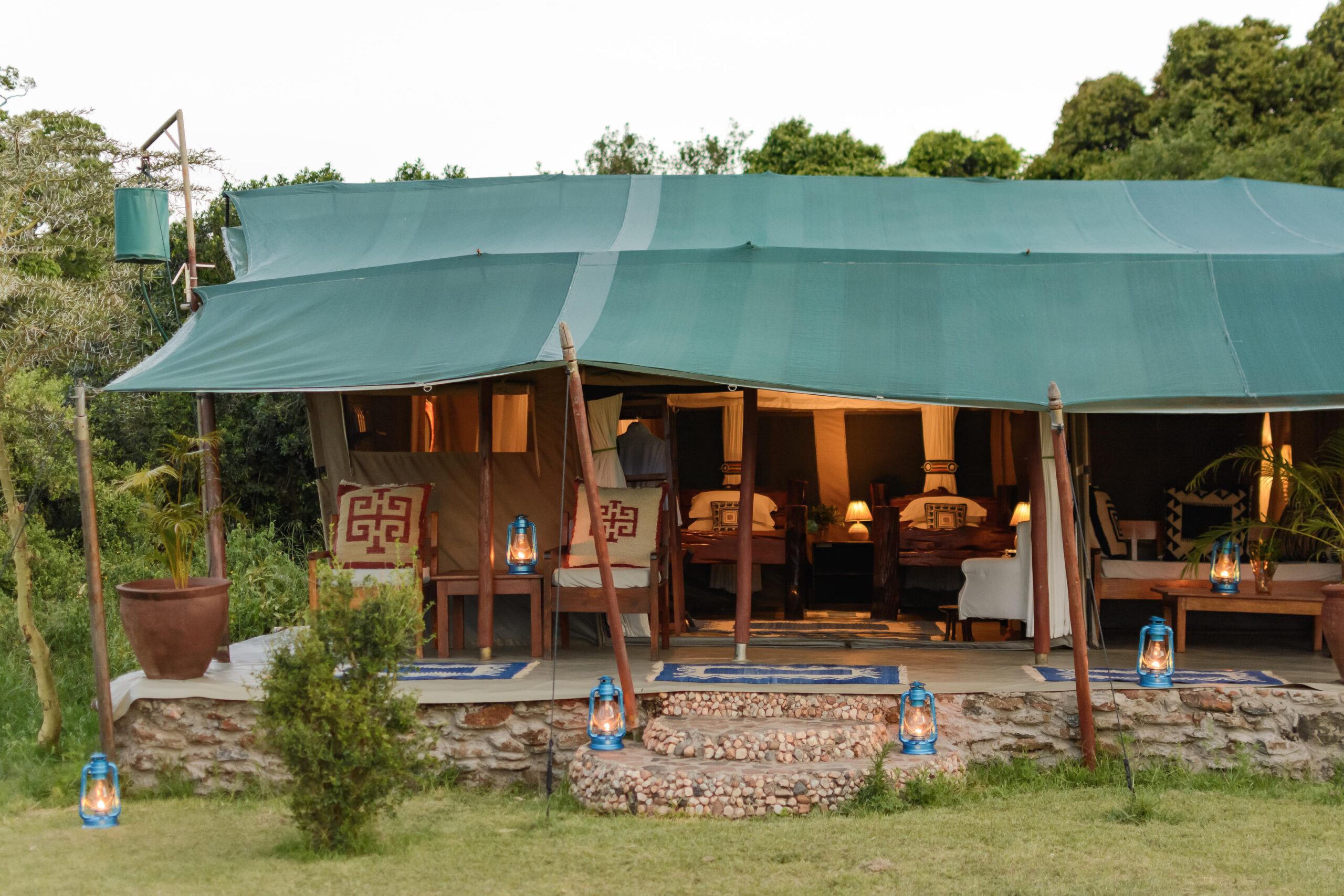 Glamping tent with a teal canvas roof, open sides, and a stone porch, featuring beds, lanterns, and wooden chairs.
