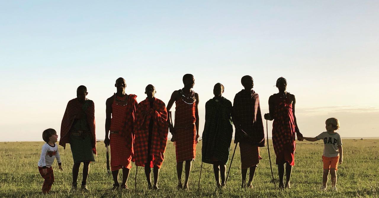 Line of Maasai adults in red and plaid shukas stands in a grassy plain at sunset, holding hands with two children at each end.