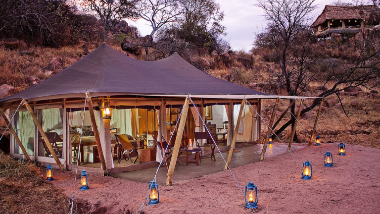 Large canvas tent pitched in a dry, rocky landscape at dusk, with wooden supports and lanterns along the perimeter.
