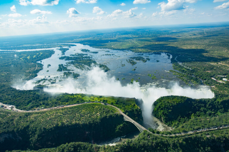 Aerial view of a wide waterfall plunging into a deep gorge with a curved bridge crossing below and lush green forests surrounding a expansive river plain.