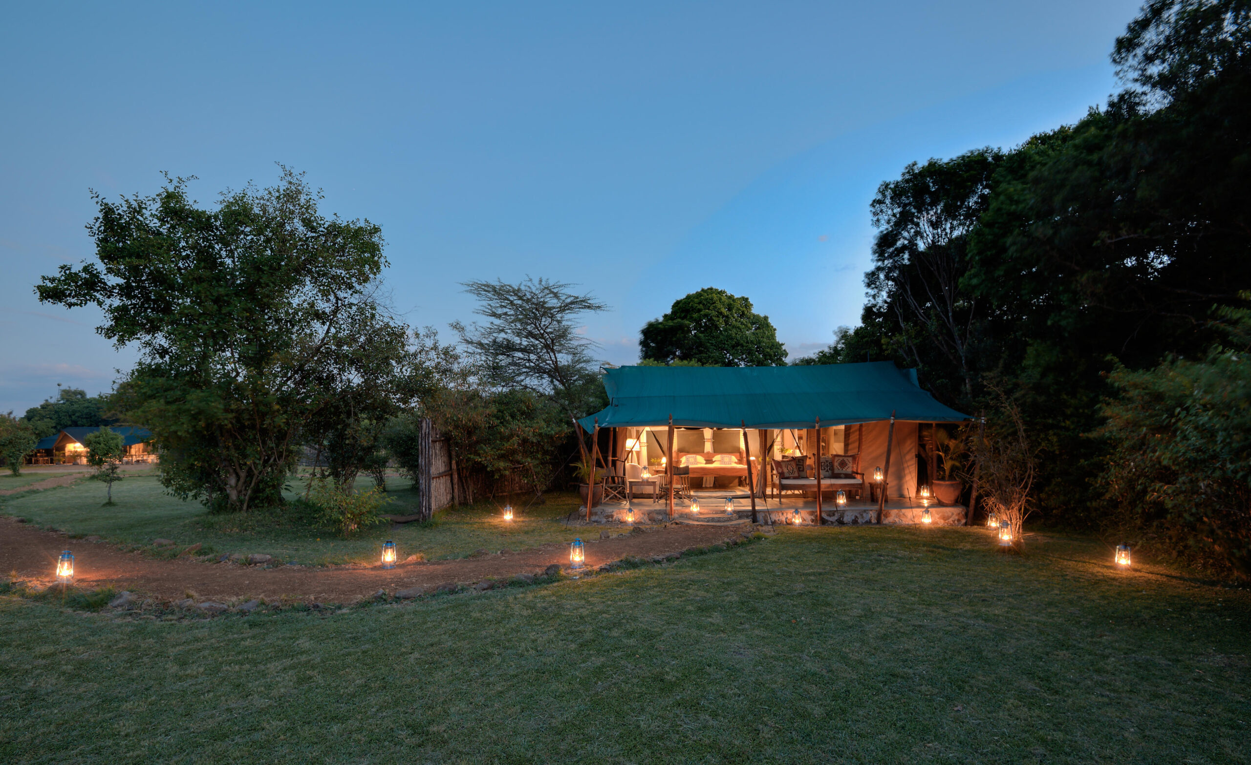 Glamping campsite with a teal-roofed tent and warm lantern light along a stone path at dusk.