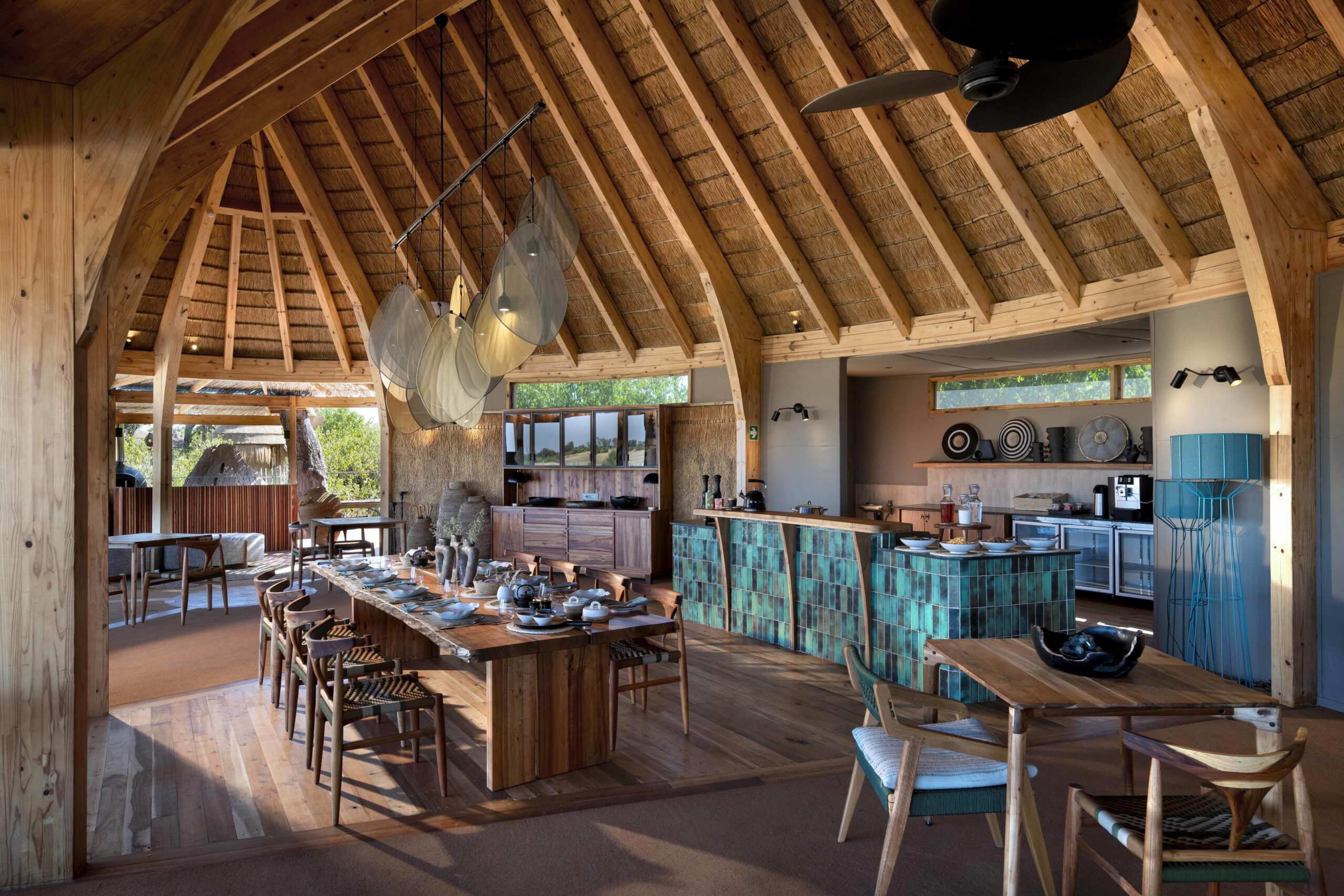 Open-plan dining area with a large wooden table set for a meal under a vaulted thatched roof and exposed wooden beams in a rustic interior.