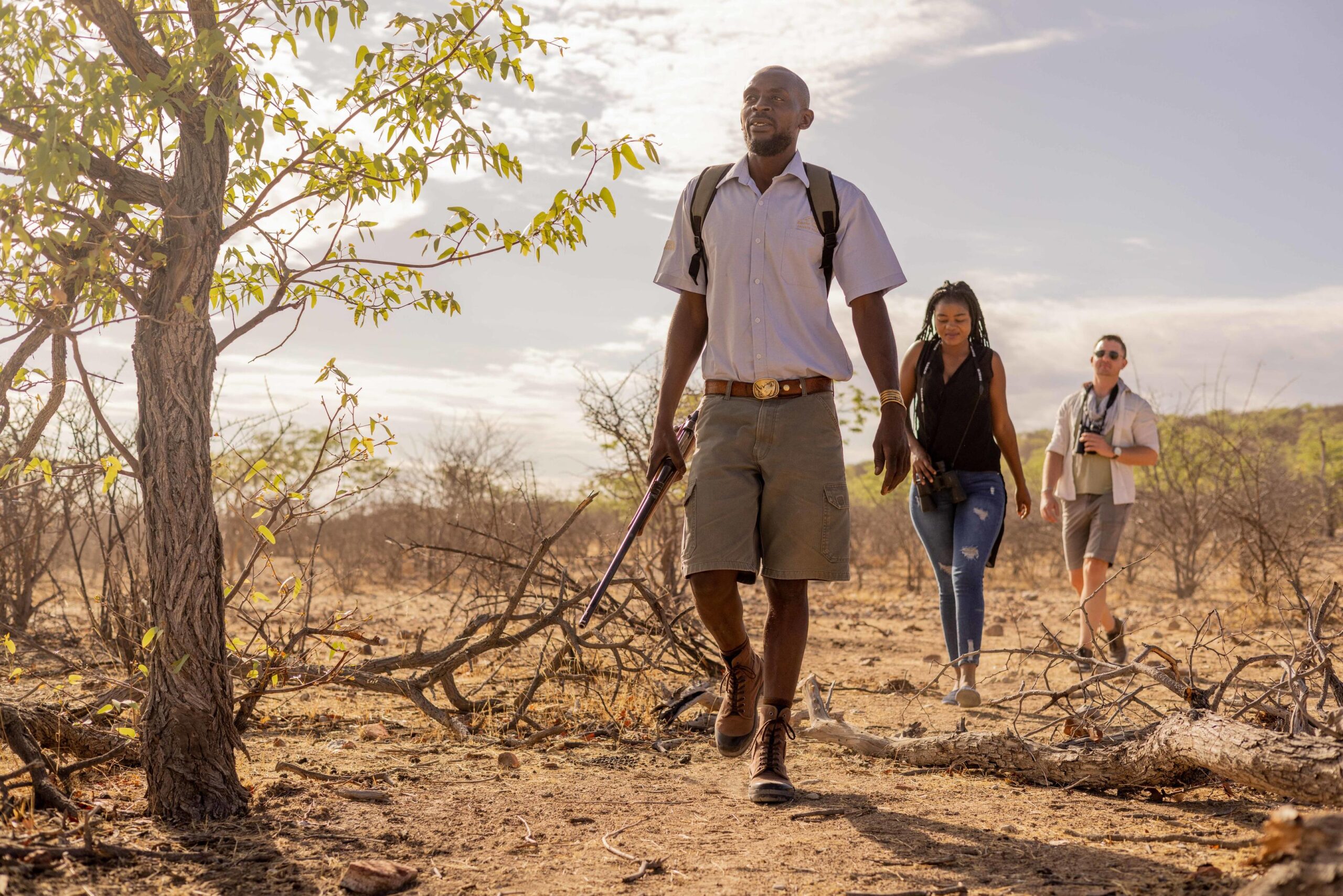 Safari guide leading two tourists across a dry savanna, holding a rifle and wearing a backpack.