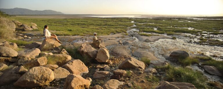 Two people sit on large rocks overlooking a tidal salt marsh at sunset with mountains in the distance.