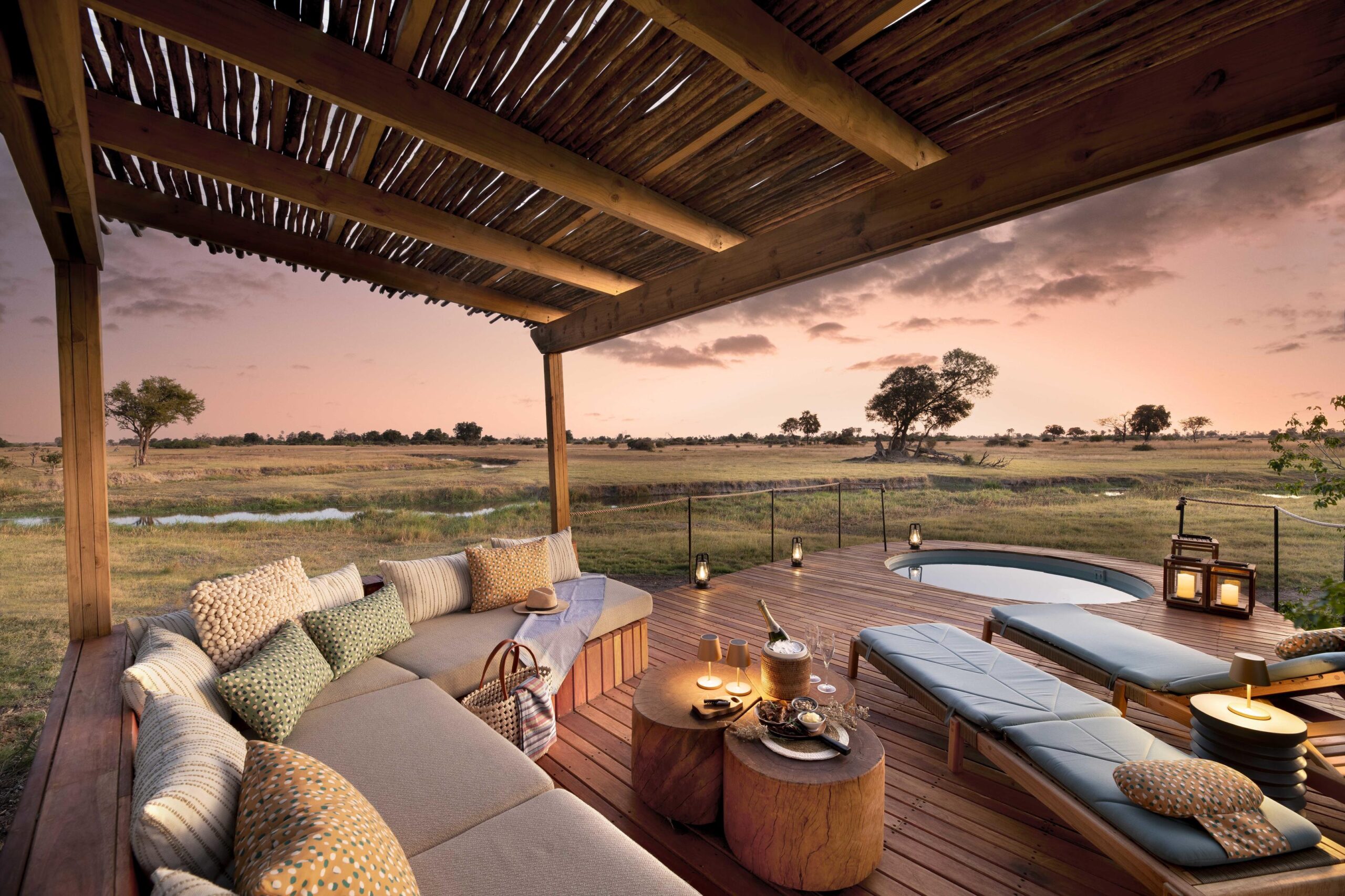Wooden deck with a cushioned seating area and round pool at sunset, overlooking a savannah with scattered trees and open grasslands.