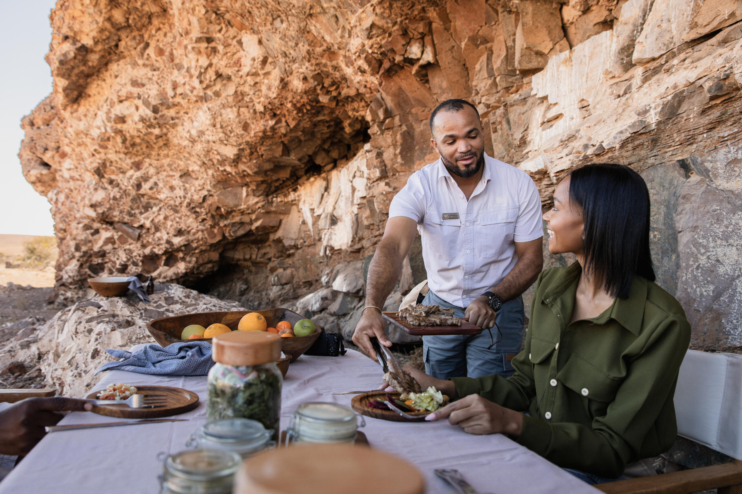 Man serving a plate of roasted meat to a smiling woman at a cliffside outdoor table with fruit bowls nearby
