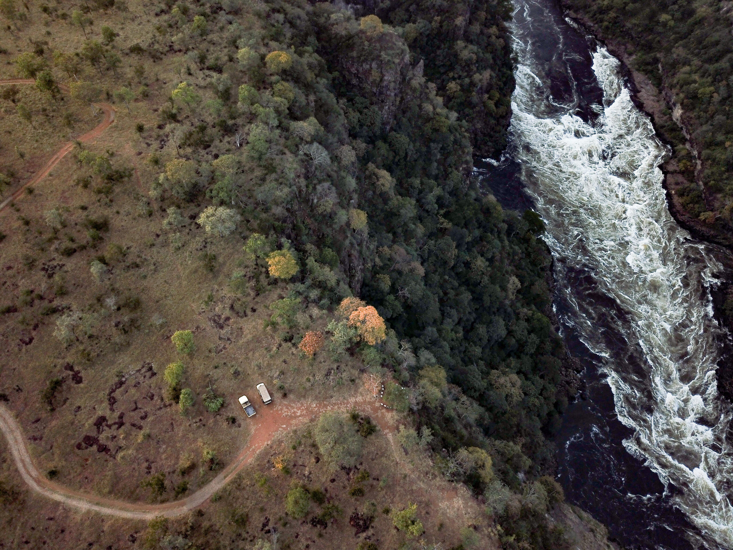 Aerial view of a winding dirt road through a grassy clearing with three parked vehicles beside trees, separated from a dense forest and a white-water river on the right