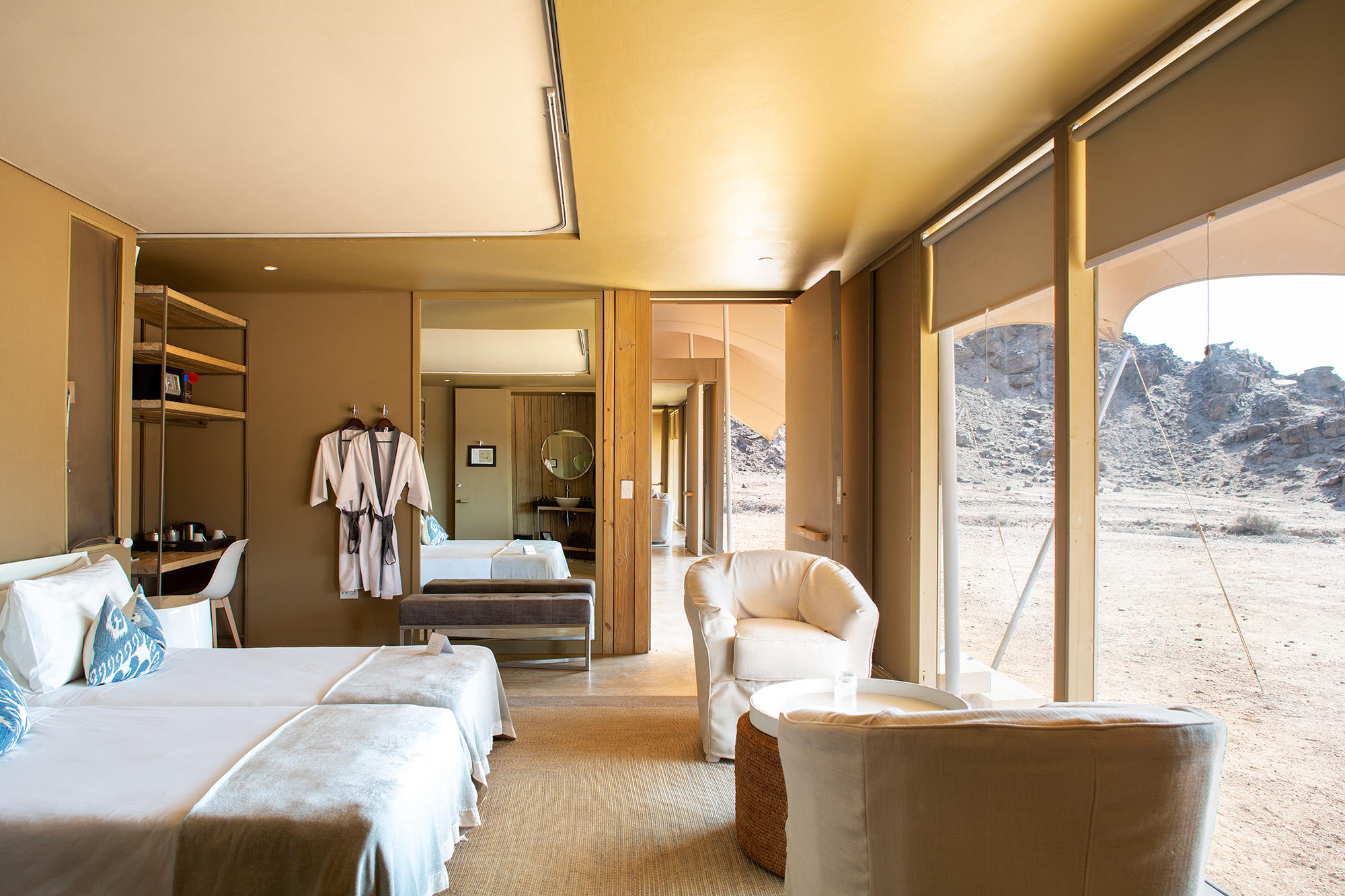 Luxurious desert-view hotel suite with two white beds, robes on hangers, and a seating area beside floor-to-ceiling windows.