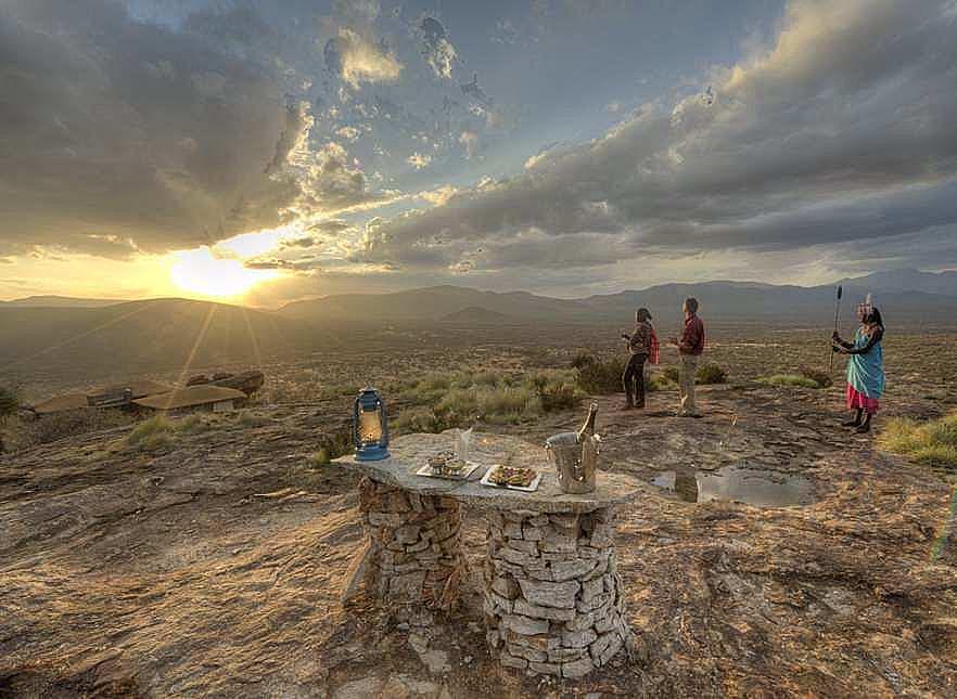 Desert sunset over a rocky plateau with a stone table set with snacks and a lantern in the foreground, and three people in colorful traditional clothing nearby.